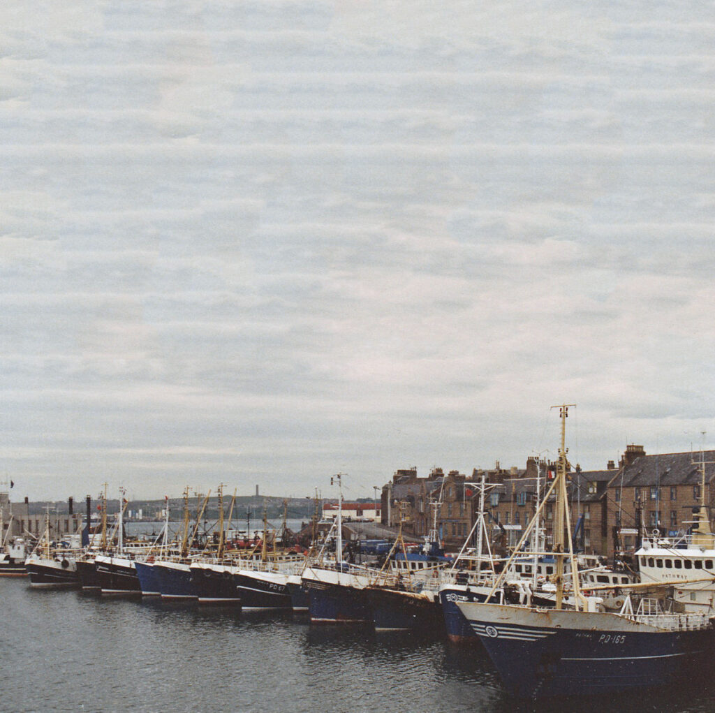 Fourteen boats lying stern-to in the south harbour, with the old lifeboat house visible in the background.